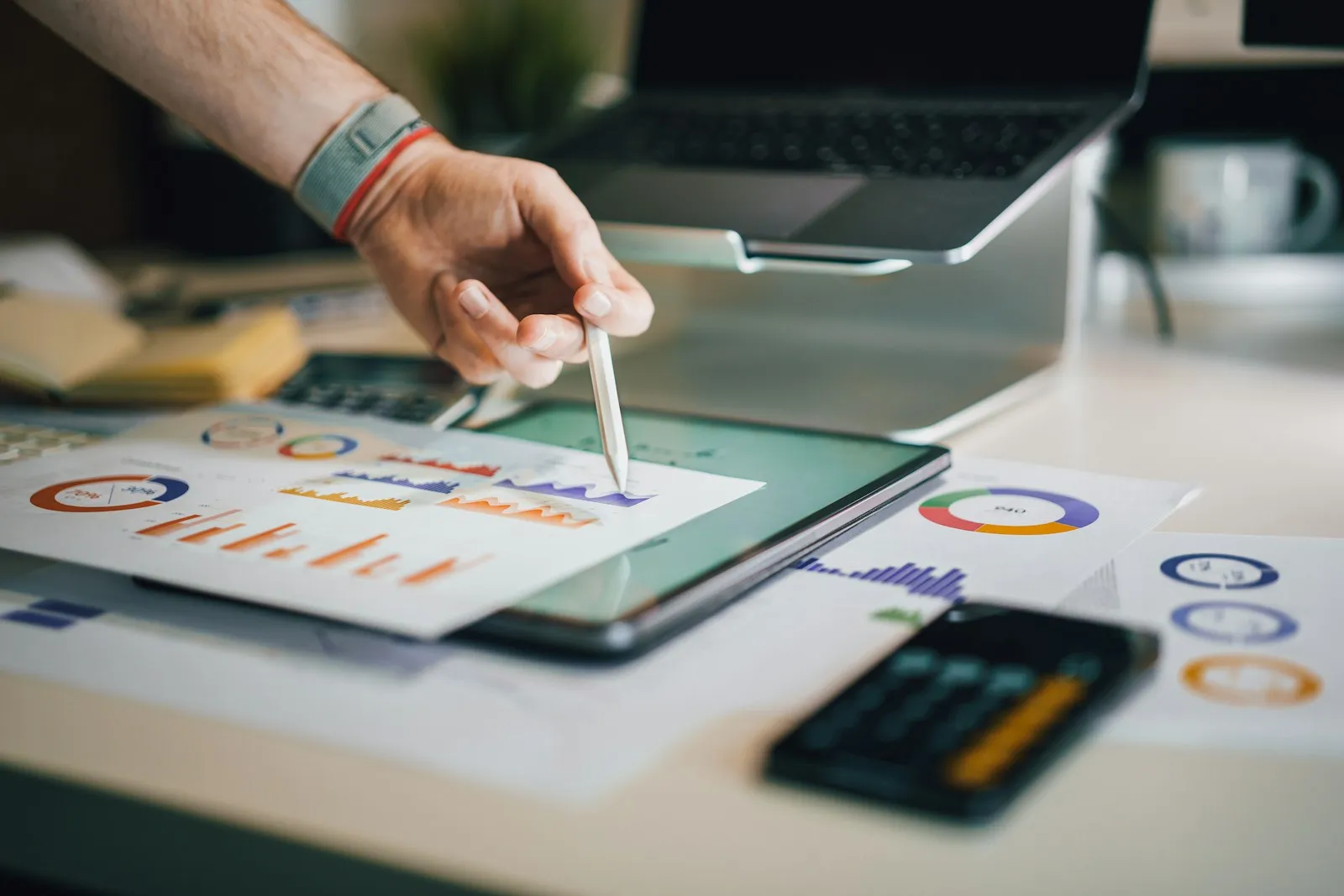 A hand pointing with a stylus at financial charts on a tablet next to printed reports
