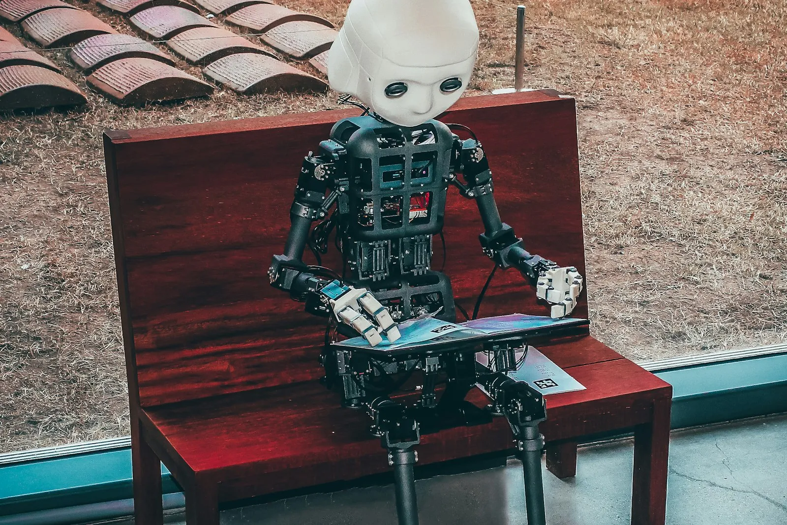 A humanoid robot seated on a bench using a laptop, as if doing a person's job