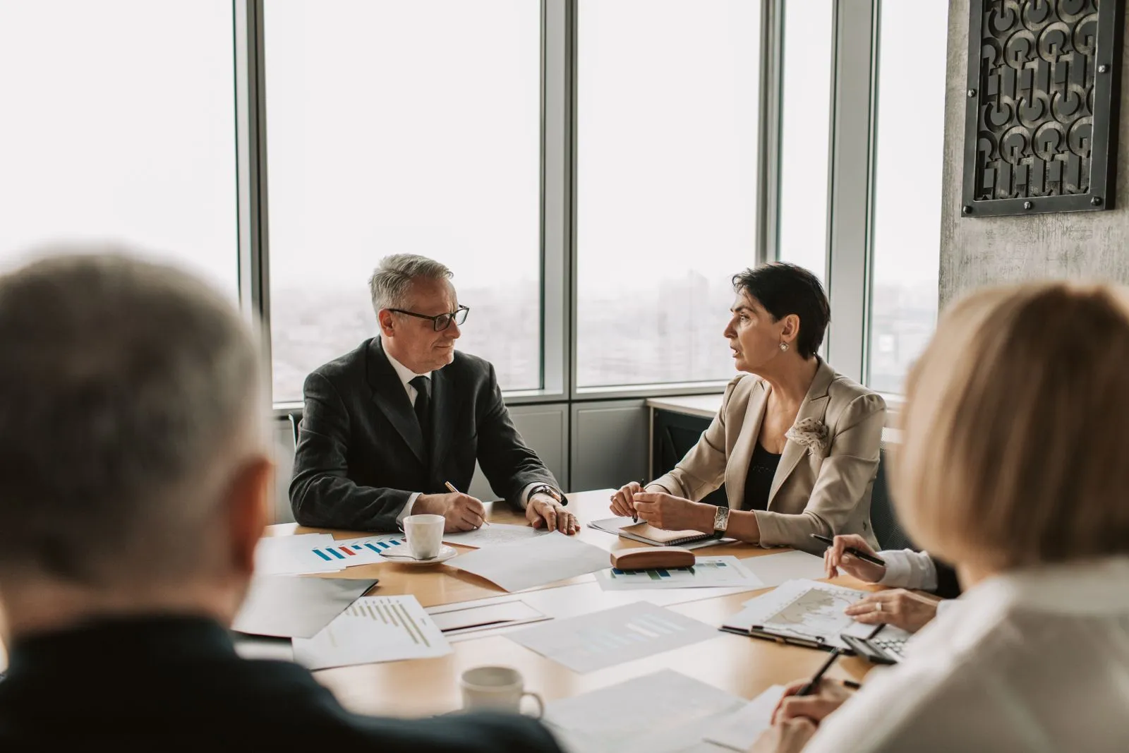 Two senior executives in conversation across a boardroom table covered with financial reports